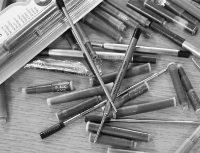 A black and white image of various pen refills and ink cartridges scattered on a wooden surface. Some are labeled with colors like Black Noir and Blue Bleu.