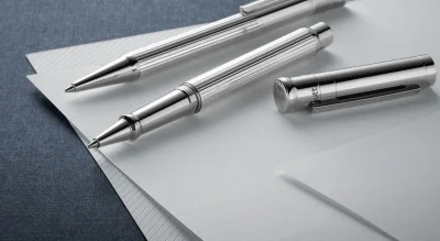 Two elegant silver pens, one capped and one uncapped, lie on top of several sheets of blank paper on a blue textured surface.
